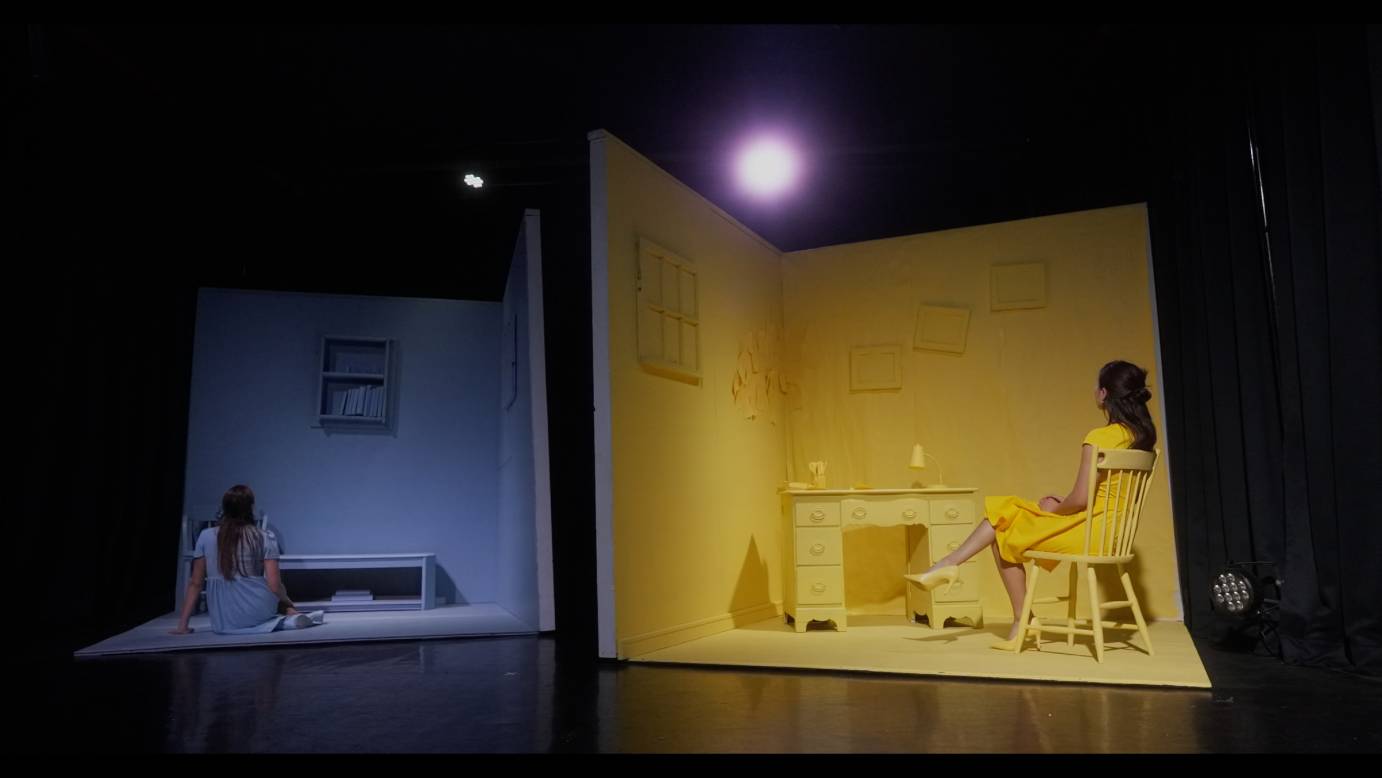a picture of two stage sets one blue and one bright yellow. Each of the rooms features a female dancer staring at the walls of her room. the yellow occupant sits formally on a chair in a proper yellow dress. the blue room features a woman sitting on the floor seeming a little more disheveled.